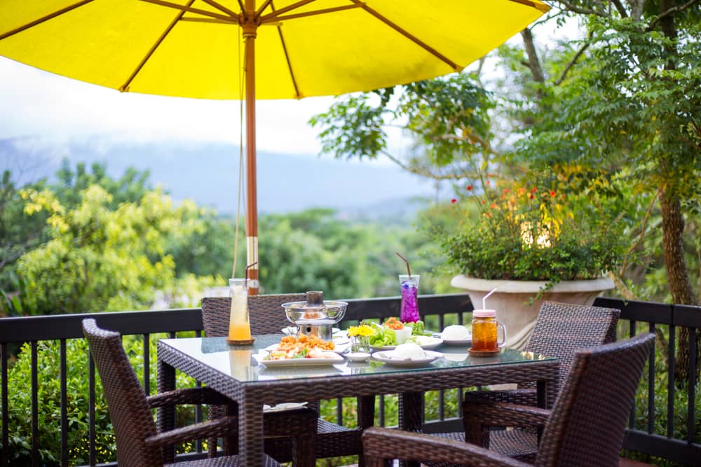 Combina los tonos neutros de las mesas de jardín con un parasol de color amarillo vibrante. Este elemento decora y complementa los exteriores.